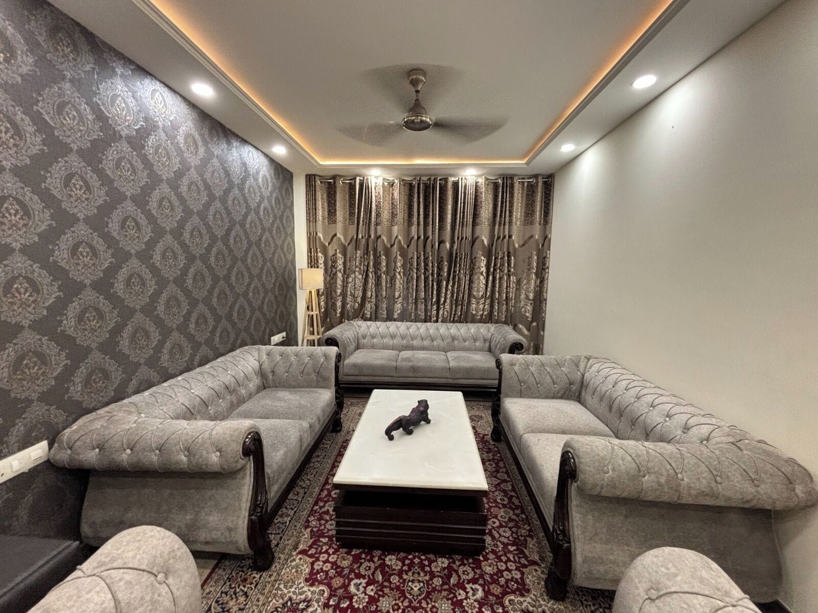 A room with a sofa set, a coffee table, patterned wallpaper, and curtains.