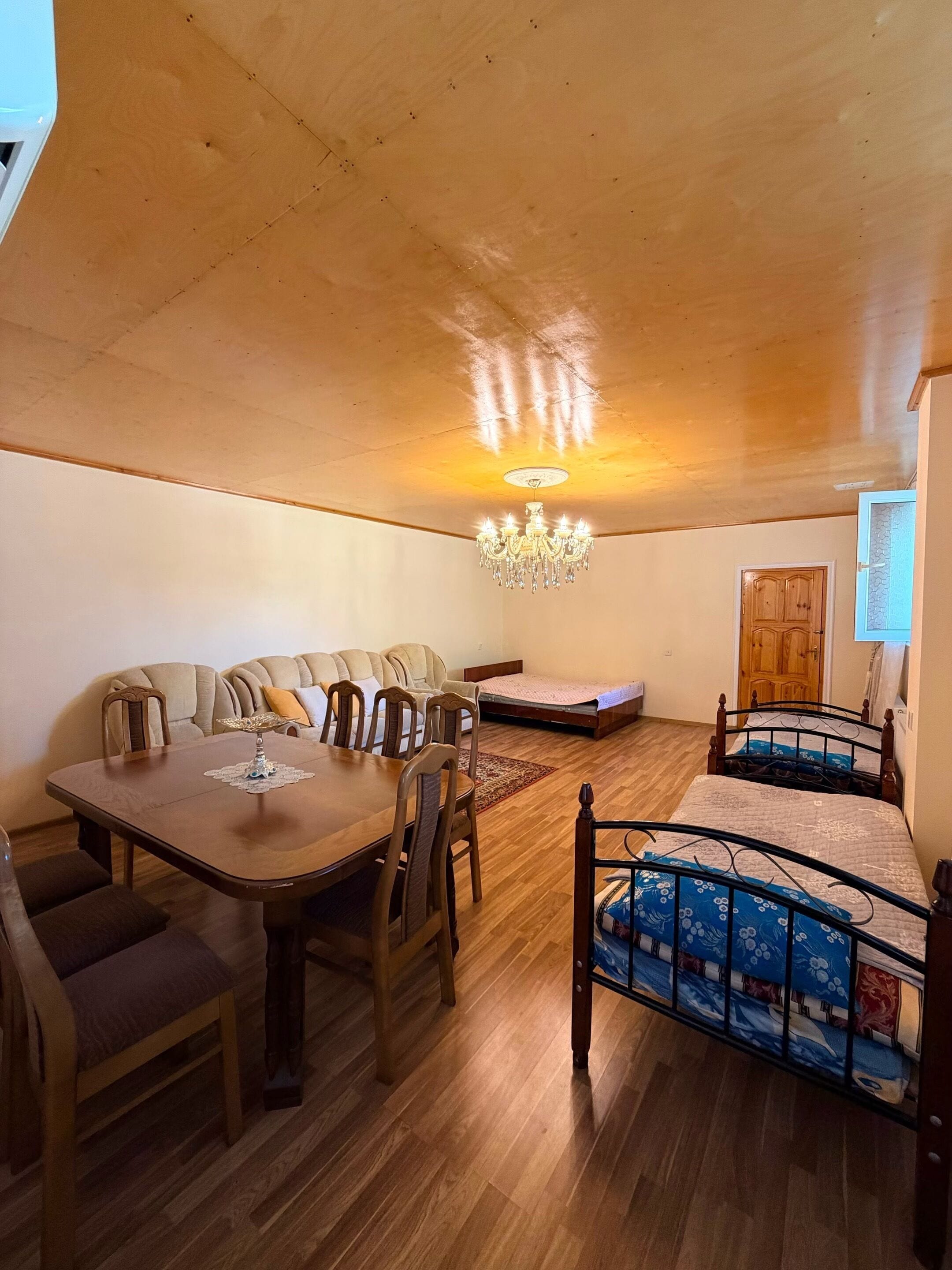 A cozy room with a wooden ceiling, a dining table with chairs, a sofa, a bed with a metal frame, and a chandelier.