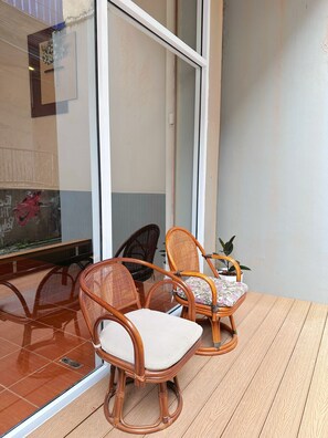 A balcony with wicker chairs and a small table, a potted plant, and a glass door reflecting a wall with a floral decoration. - Green Hill Hostel (Vientiane)