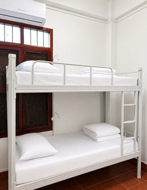 Un lit superposé avec une literie blanche, une fenêtre avec un cadre en bois et un appareil de climatisation installé au-dessus.