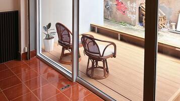 A room with a large sliding glass door leading to an outdoor area with a view of a blue building and a mural. There are two wicker chairs and a potted plant inside.