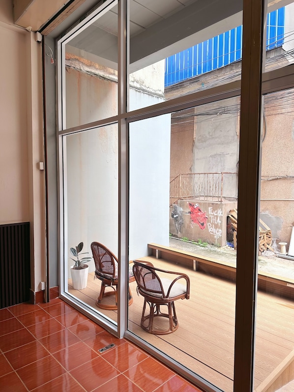 A room with a large sliding glass door leading to an outdoor area with a view of a blue building and a mural. There are two wicker chairs and a potted plant inside.