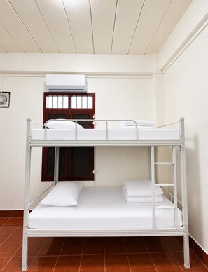 Una cama litera con ropa de cama blanca en una habitación con piso de baldosas y ventana con marco de madera.