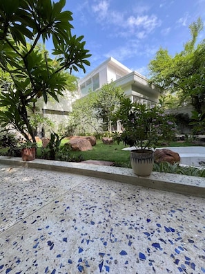 Uma passarela pavimentada com plantas em vasos, um edifício com amplas janelas e uma área gramada com pedras.