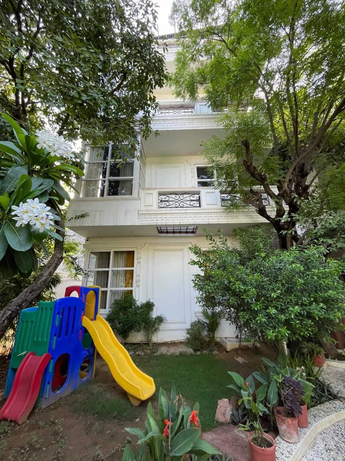 Un edificio de dos pisos con fachada blanca, un balcón con barandilla decorativa y un pequeño jardín con un tobogán y plantas en macetas.
