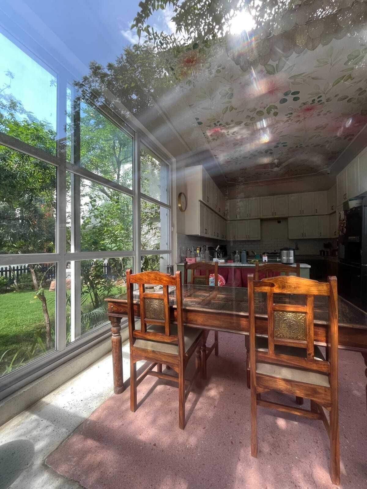 A dining area with a glass table and wooden chairs, a kitchen in the background, and a view of a garden through large windows.