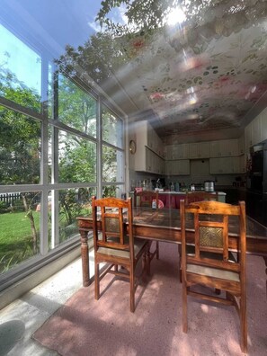 Uma sala de jantar com mesa de vidro e cadeiras de madeira, cozinha ao fundo e vista para um jardim através de grandes janelas.