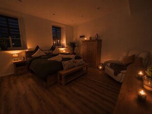 Une chambre à coucher faiblement éclairée, avec un grand lit, des tables de chevet, une armoire en bois et un coin salon avec un fauteuil.
