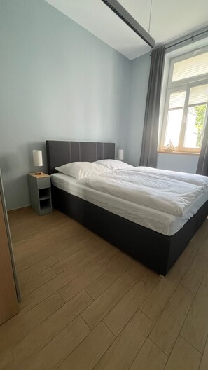 A modern bedroom with a large bed, a bedside table, and a window with curtains. - Vrbo Property (Rostock)