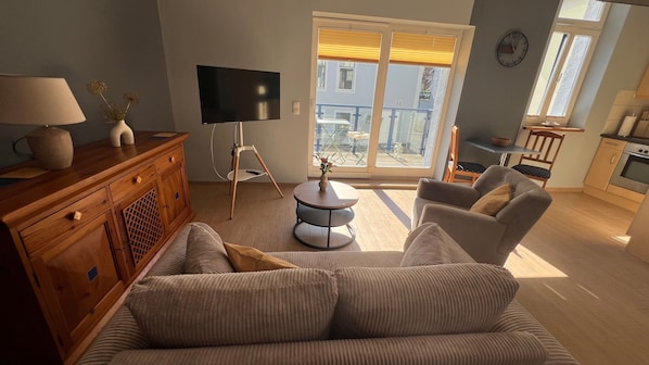 A living room with a sofa, a TV, a wooden cabinet, and a small round table with a vase. - Vrbo Property (Rostock)