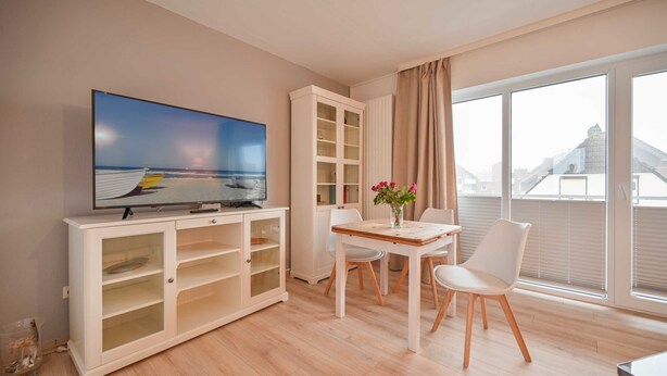 A modern living room with a flat-screen TV, a dining area with a table and chairs, a white cabinet, and large windows with curtains. - Vrbo Property (Kellenhusen)