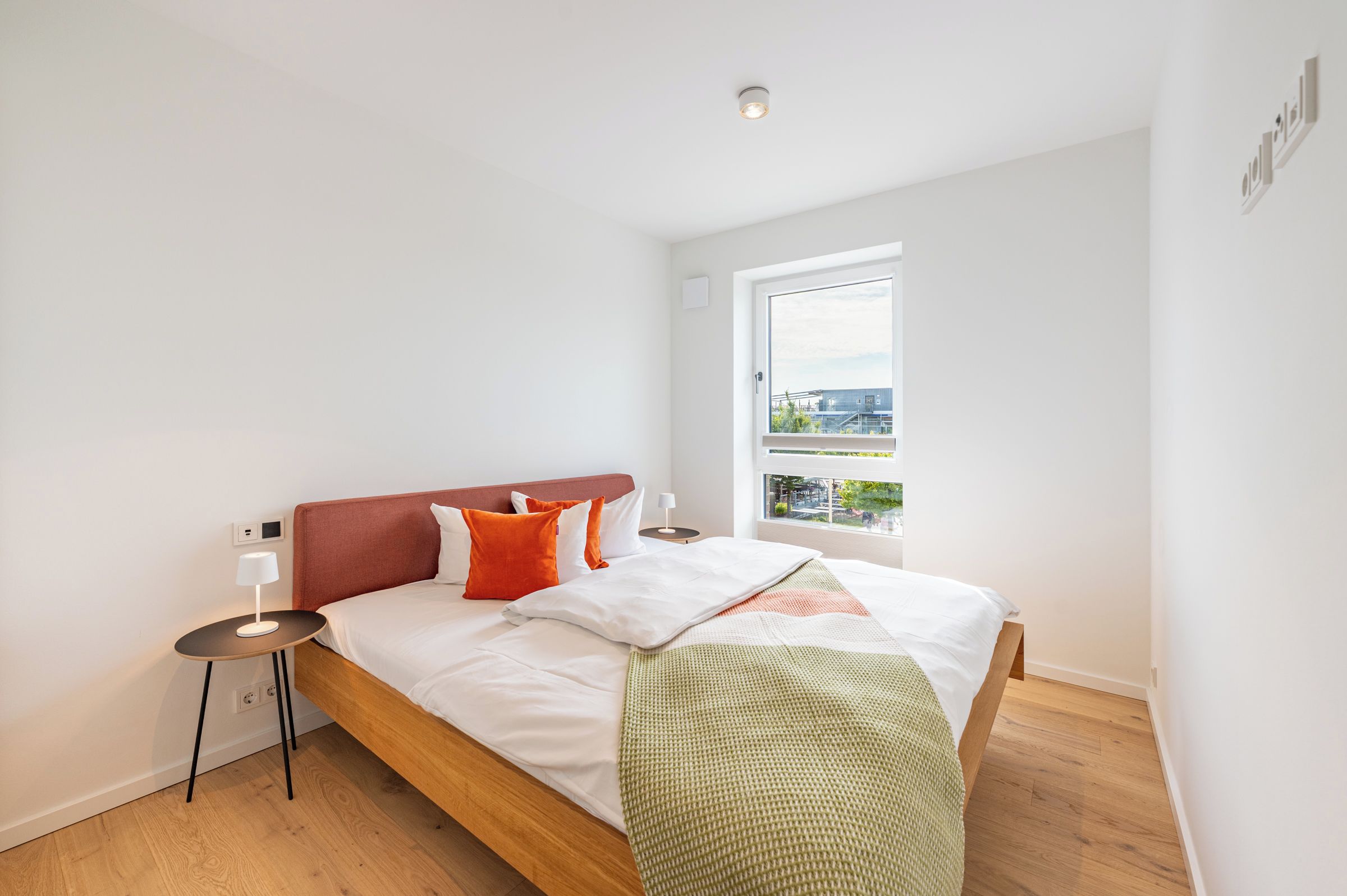 A modern bedroom with a wooden bed, white bedding, and a view of an industrial area through a large window.
