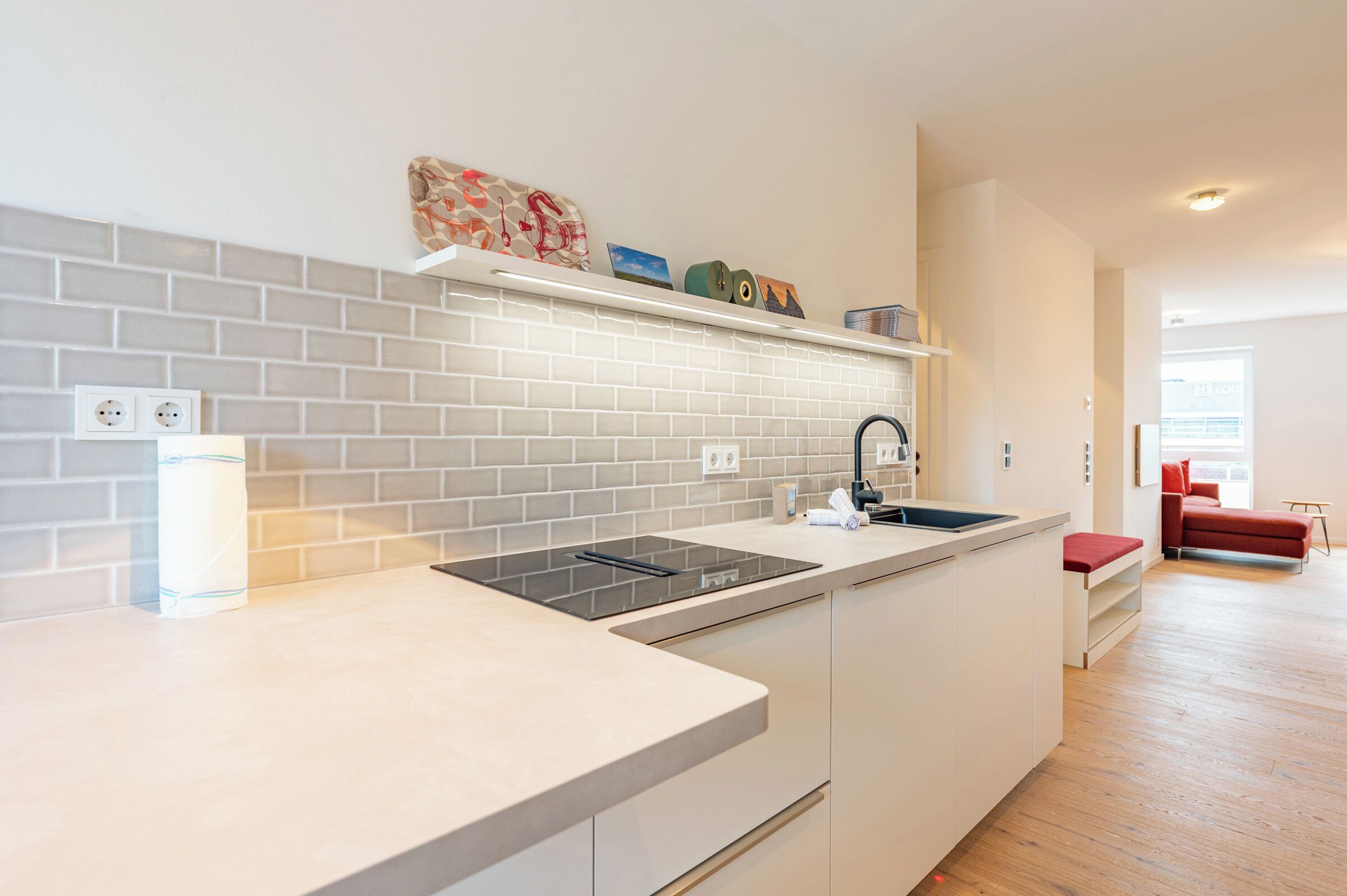 A modern kitchen with a white countertop, a built-in stove, and a sink. There is a shelf above the counter with decorative items.