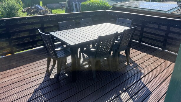 Eine Terrasse im Freien mit Tisch und Stühlen, umgeben von einem Holzzaun, mit Blick auf einen Garten und Solaranlagen.