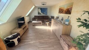 Ein moderner Wohnraum mit schrägem Dach, einem Fernseher, einer Couch, einem Couchtisch und einer Küchenzeile mit Arbeitsplatte und Schränken.