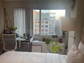 Ein Hotelzimmer mit Bett, Schreibtisch, Stuhl, einem Fenster mit Jalousien und einigen Kübelpflanzen.