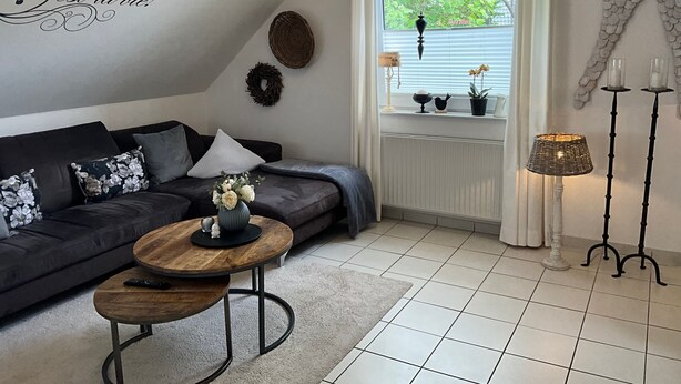 A modern living room with a dark sofa, a round wooden coffee table, and a window with a view of greenery. - Vrbo Property (Cloppenburg)