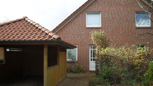 A brick house with a tiled roof, a wooden structure to the left, and a garden area with bushes and a tree. - Vrbo Property (Cloppenburg)