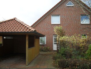 A brick house with a tiled roof, a wooden structure to the left, and a garden area with bushes and a tree. - Vrbo Property (Cloppenburg)