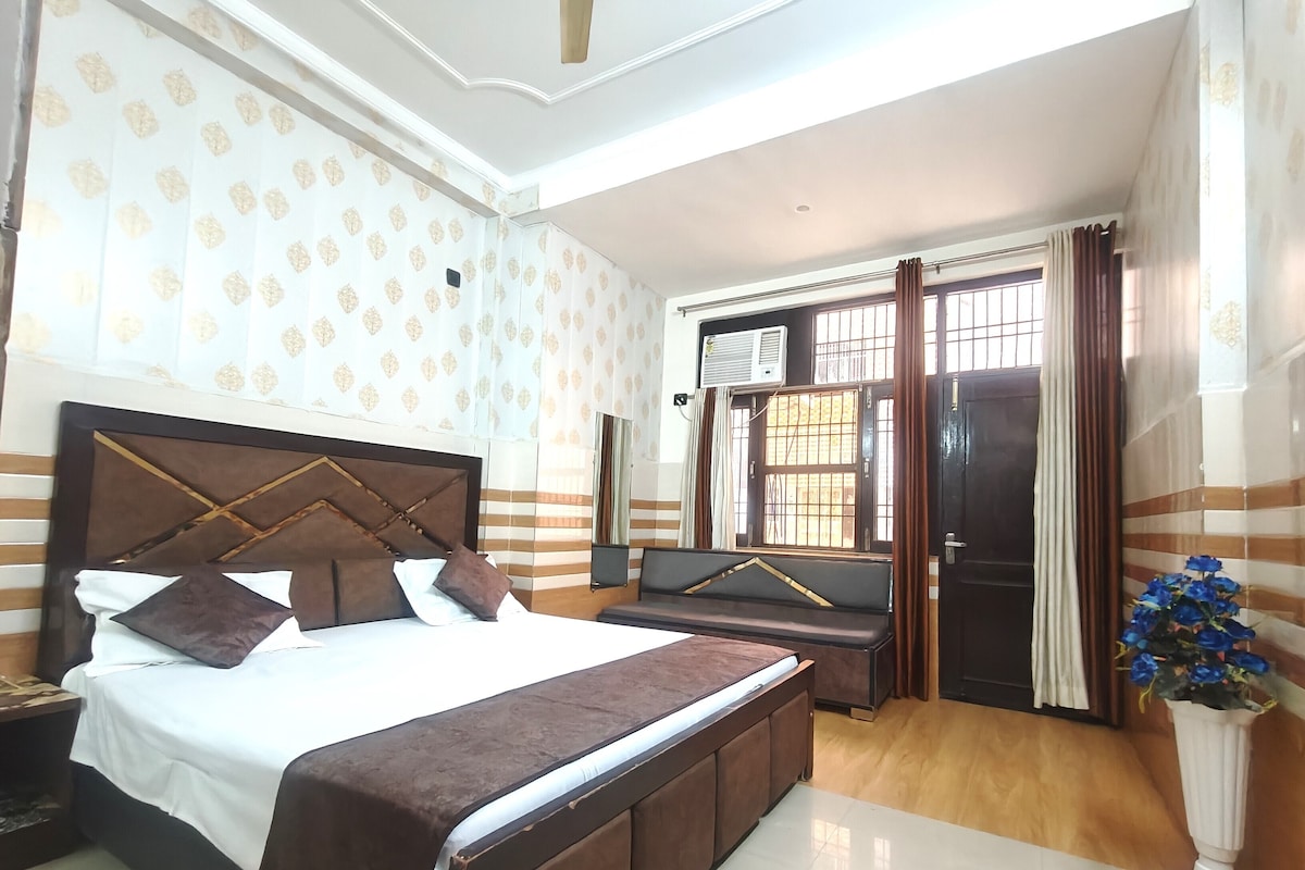 A hotel room with a bed, a seating area, a window with curtains, and a potted plant.