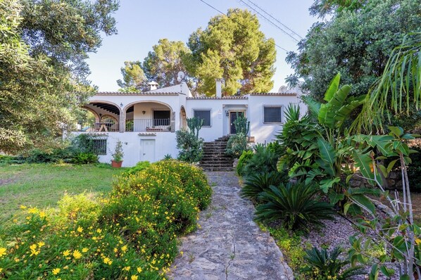 A white building with a stone pathway leading to the entrance, surrounded by lush greenery and flowering plants. - Vrbo Property (VC)