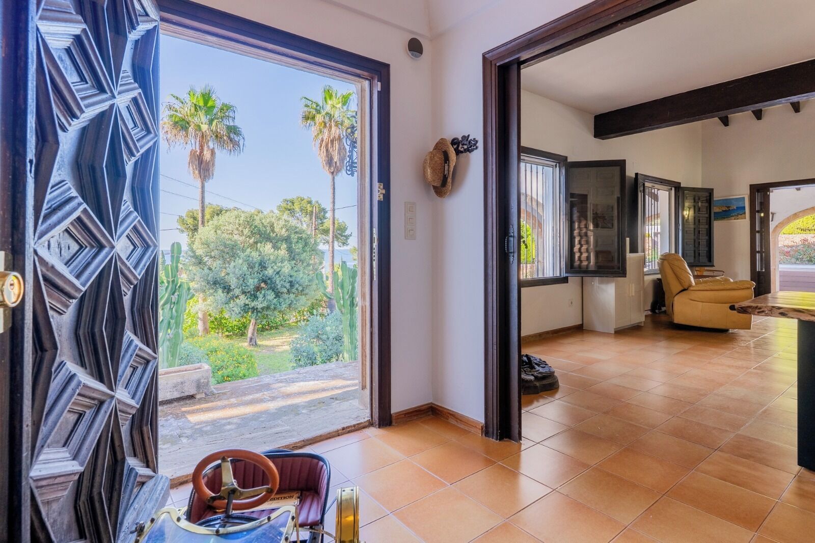 A spacious room with a large sliding glass door leading to a garden, a comfortable seating area with a sofa and armchairs, and a kitchen area visible through the door.
