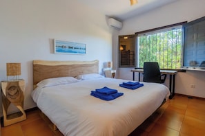 A hotel room with a bed, a desk, a chair, a window with a view of greenery, and a painting on the wall.