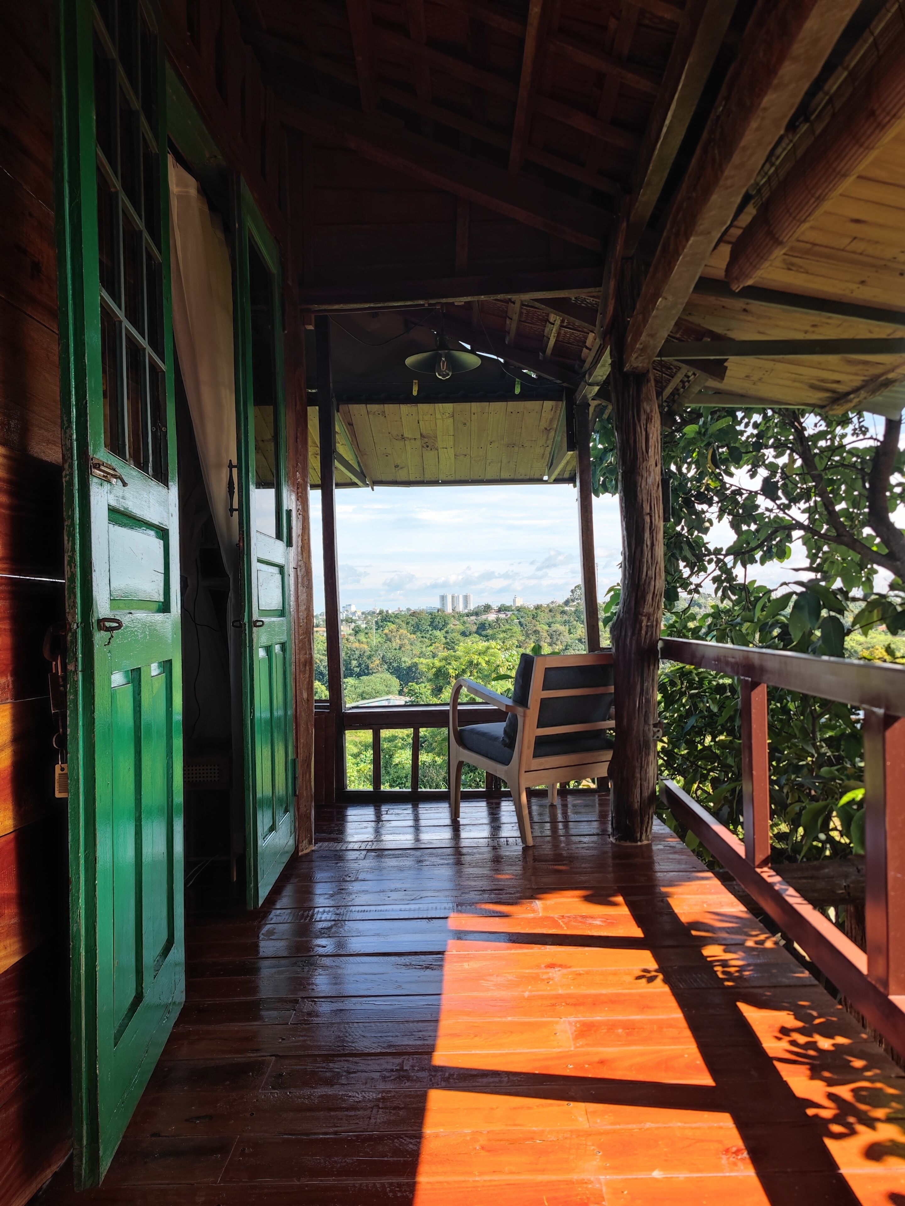 Un porche de madera con un banco, vista a un paisaje urbano y una puerta verde.