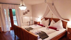 Llits dobles amb estructura de fusta, teles de llit blanques i un dosser. Hi ha una taula de nit amb una làmpada, una petita estanteria amb làmpada i un armari petit amb nansa en forma de cor.