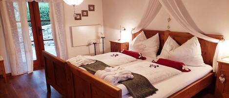 A double bed with a wooden frame, white linens, and a canopy. There is a bedside table with a lamp, a nightstand with a lamp, and a small cabinet with a heart-shaped handle.