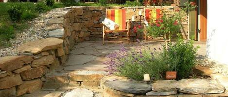 A stone pathway leading to a seating area with a chair and a table, surrounded by plants and a stone wall.