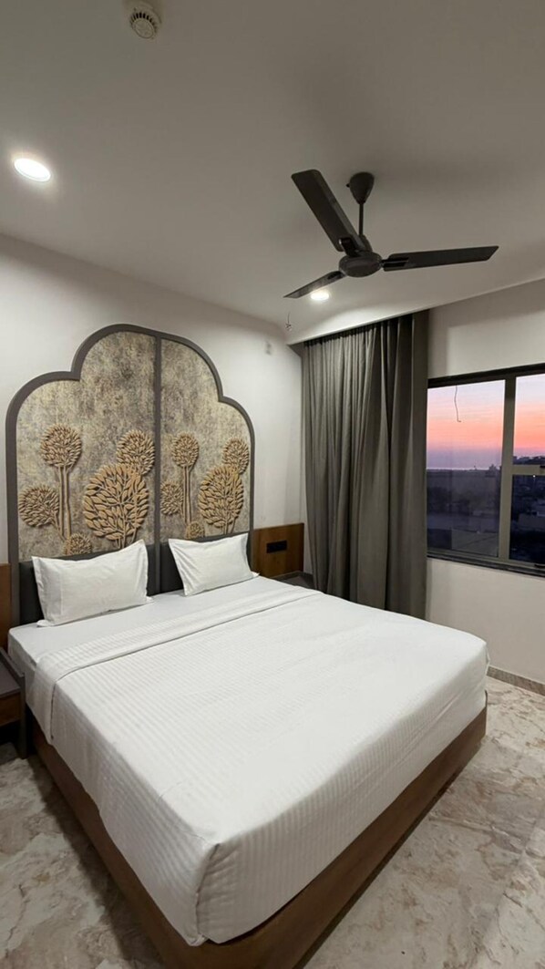 A bedroom with a large bed, a ceiling fan, a nightstand, a window with curtains, and a view of the cityscape. - Hotel Ishant (Dwarka)
