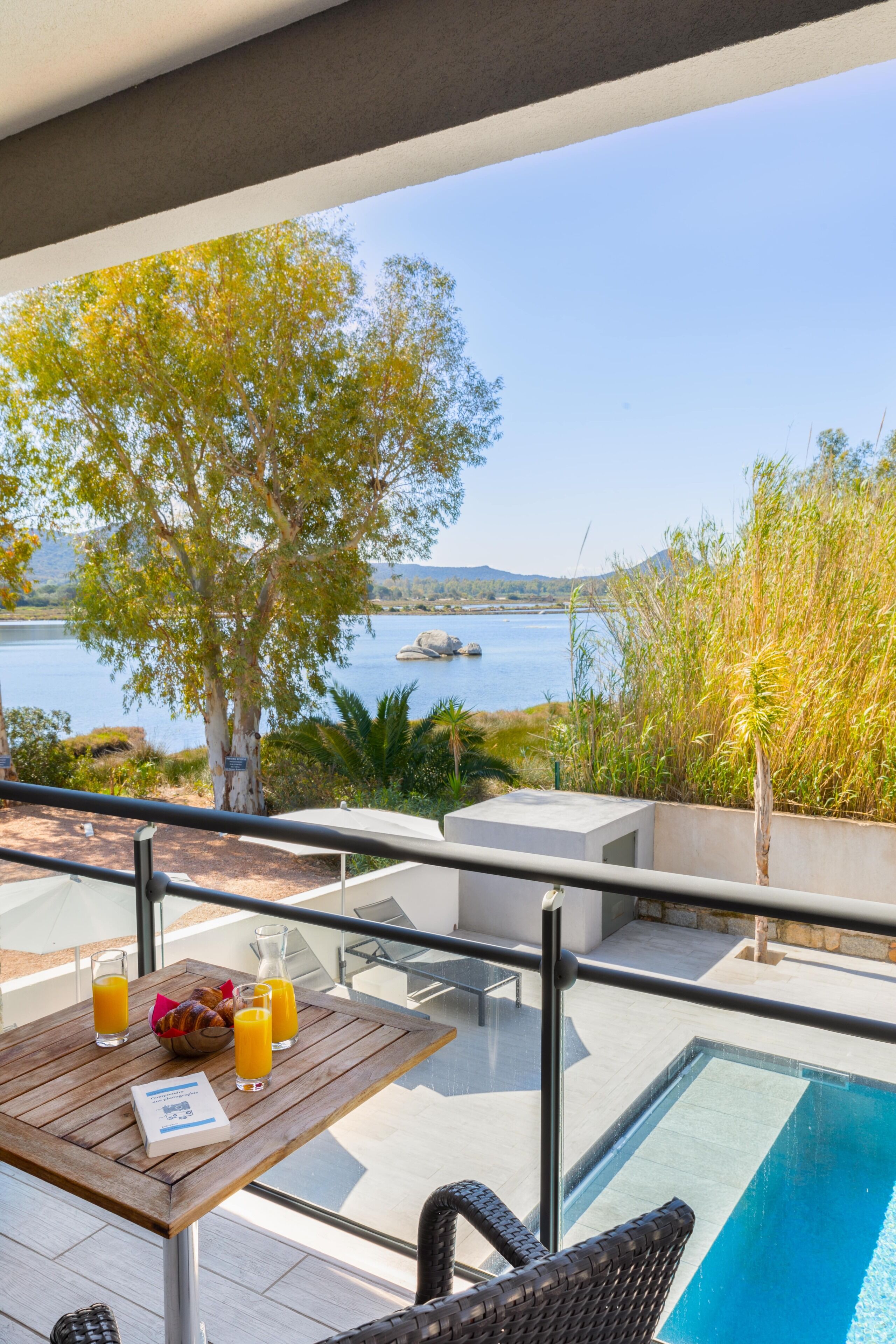 Un balcon avec une table préparée pour le petit-déjeuner, donnant sur une étendue d’eau et un arbre.