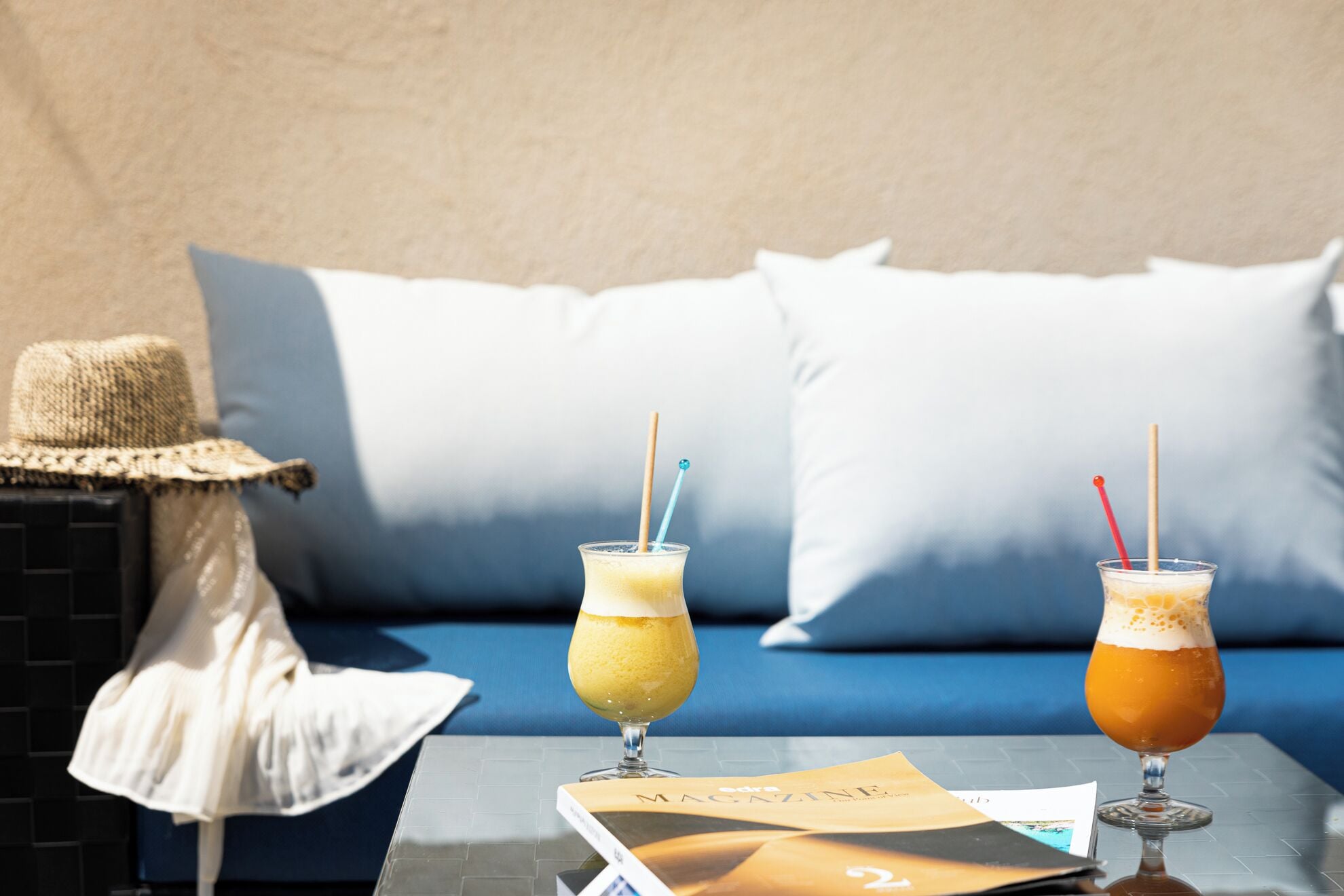 A table with two drinks, a straw, a magazine, and a straw hat.