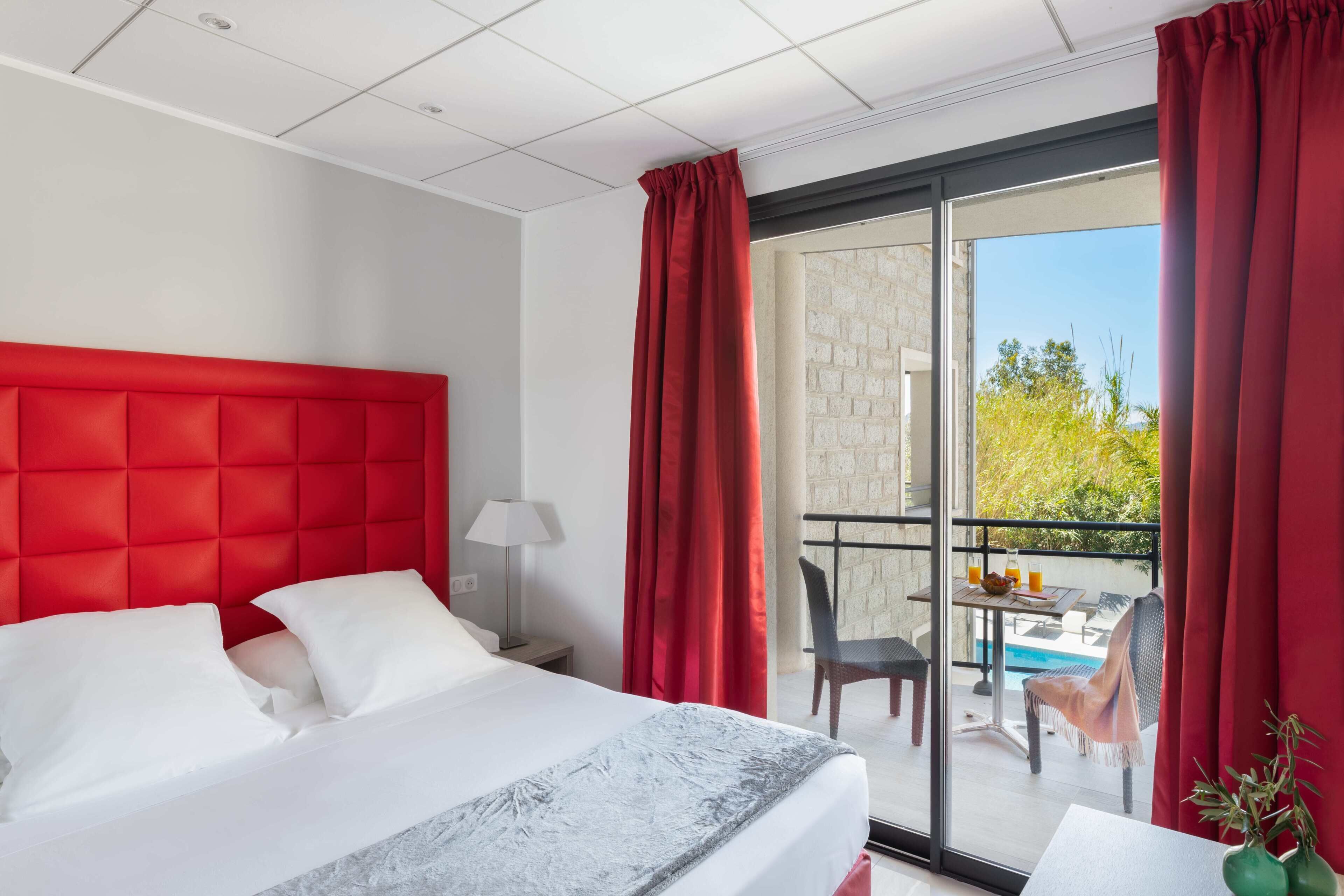 Une chambre d’hôtel avec un grand lit, un bureau, une chaise, une table avec une boisson, et une vue sur l’extérieur.