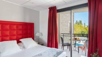 Une chambre d’hôtel avec un grand lit, un bureau, une chaise, une table avec une boisson, et une vue sur l’extérieur.