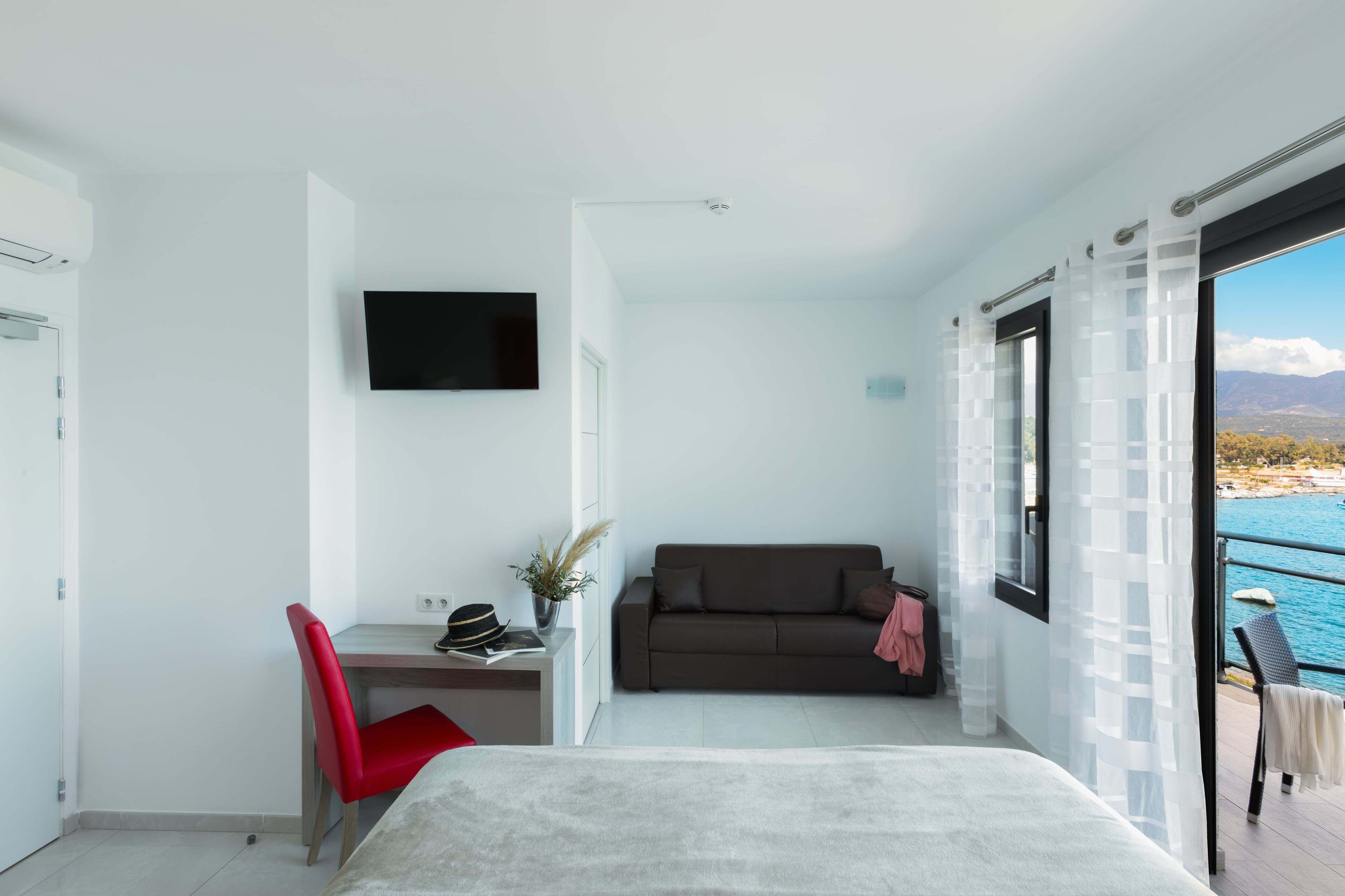 Une chambre moderne avec un lit, un bureau, un fauteuil rouge, une télévision, un canapé et une vue sur la mer.