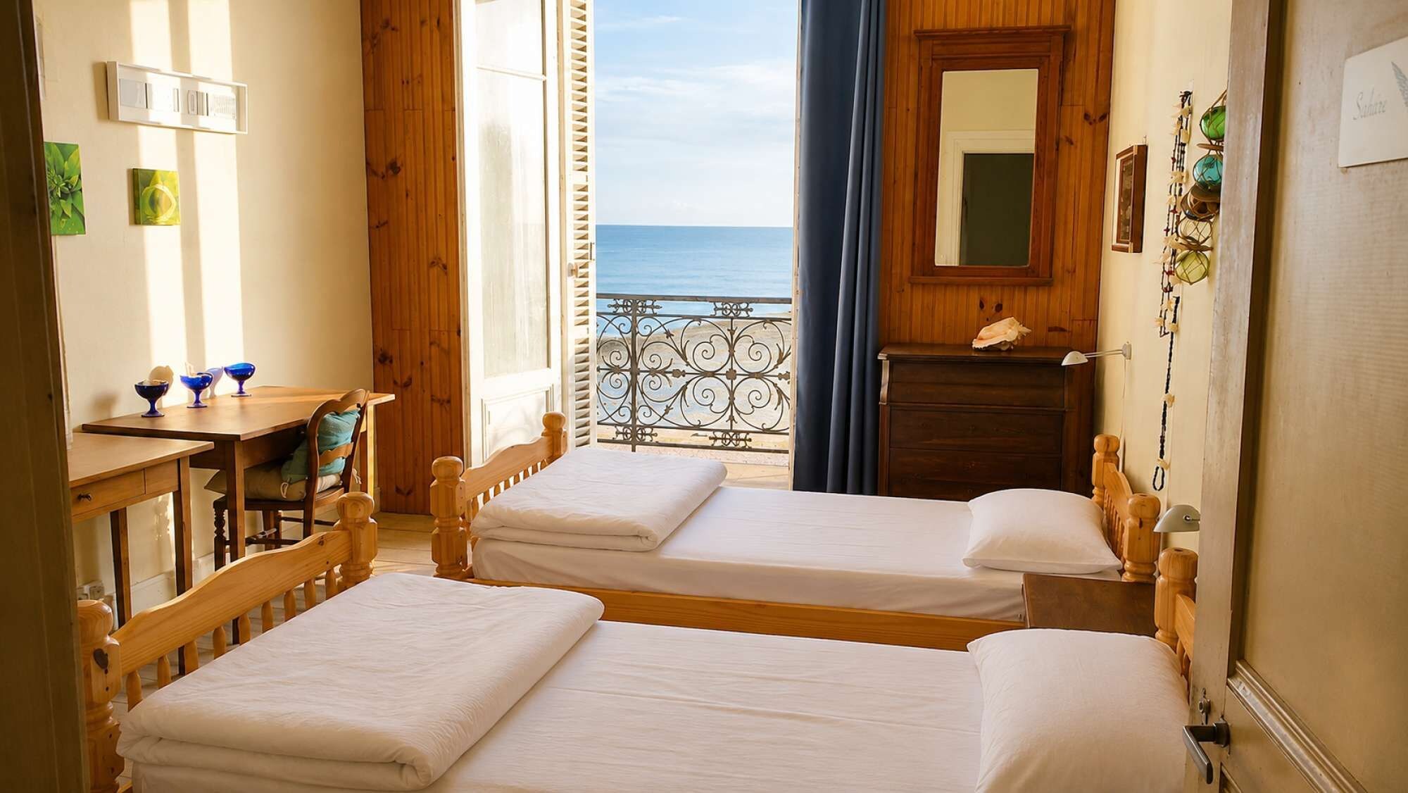 A room with two beds, a wooden desk, a dresser, and a view of the sea.
