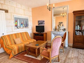 A furnished living room with a sofa, a television, a wooden coffee table, a chair, a bookshelf, and a mirror. - Villa Anne-Marie - 19th-century house with ocean views (Saint-Aubin-sur-Mer)