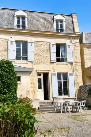 Un bâtiment à deux étages, doté d’une façade en pierre, de nombreuses fenêtres avec volets et d’un petit espace extérieur aménagé avec des chaises en plastique.