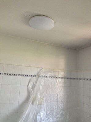 A room with white tiled walls, a ceiling with a round light fixture, and a plastic sheet covering part of the wall. - Breakfast and Beverages (Biella)
