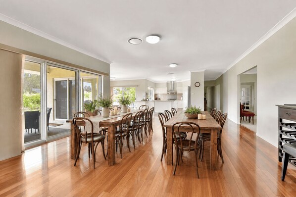 A spacious dining area with a long wooden table and chairs, a kitchen in the background, and a sliding glass door leading to an outdoor area.