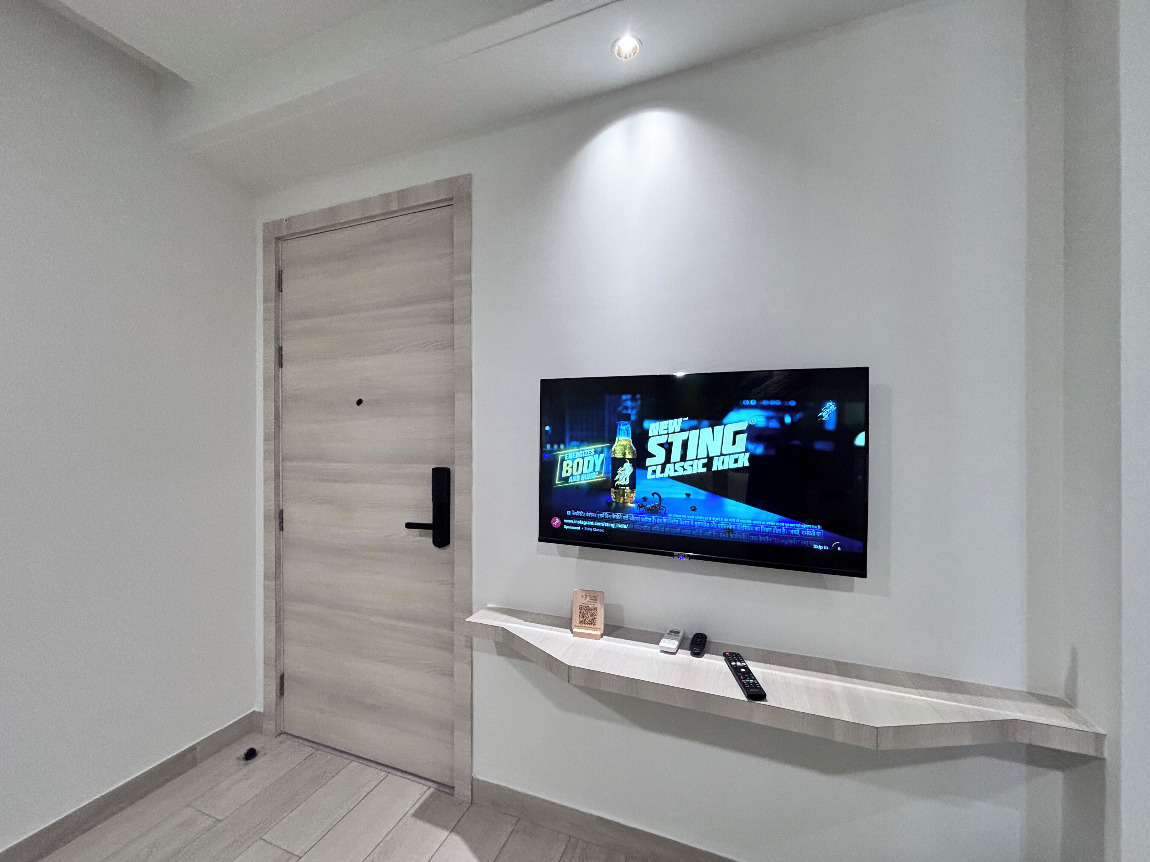 A modern hallway with a door, a mounted TV displaying an advertisement, and a shelf with a remote control.