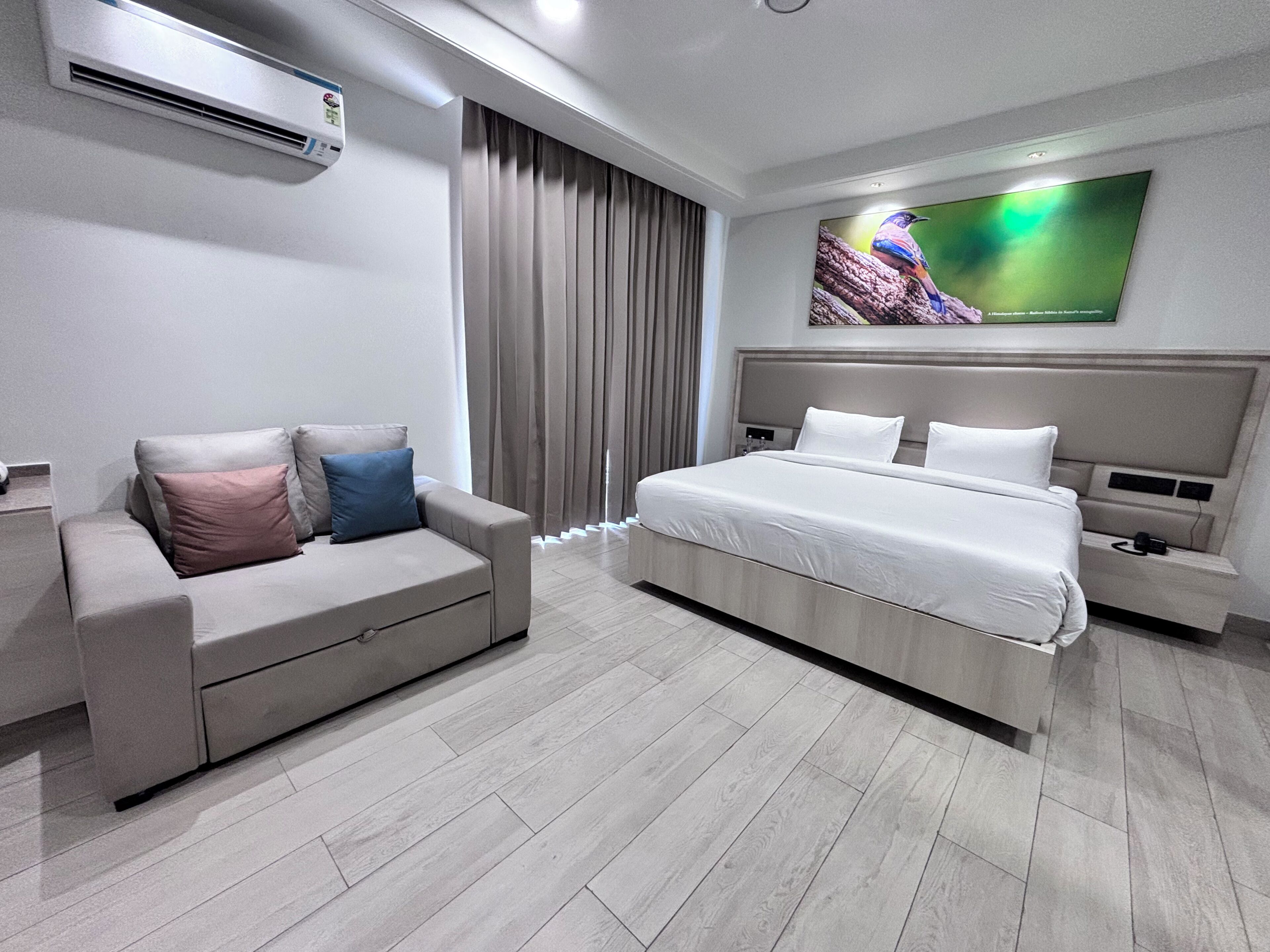 A modern hotel room with a bed, a sofa, a TV, and an air conditioning unit.