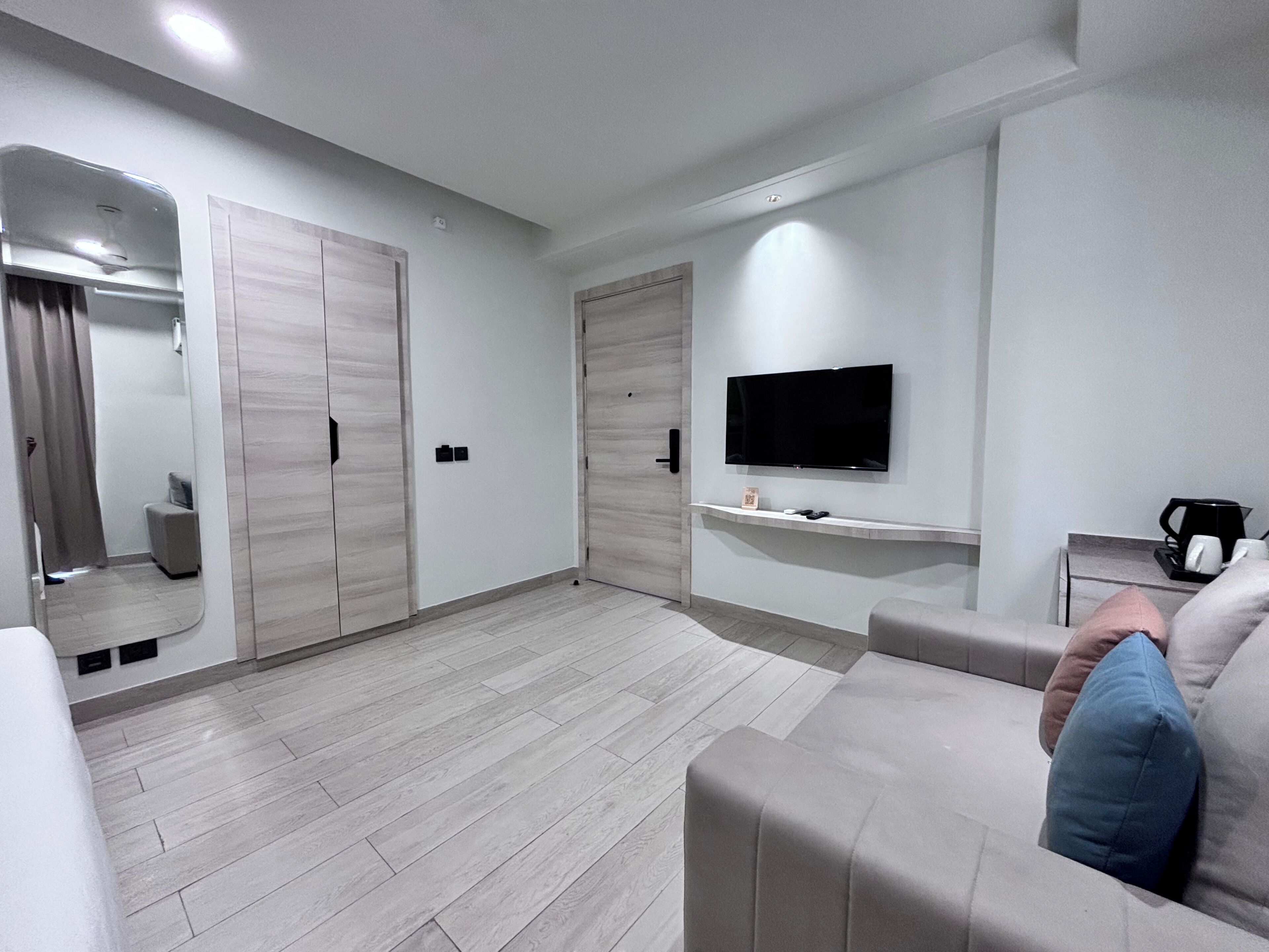 A modern living room with a flat-screen TV mounted on the wall, a sofa with cushions, a coffee machine, and a small table.