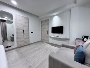 Ein modernes Wohnzimmer mit einem wandmontierten Flachbildfernseher, einer Couch mit Kissen, einer Kaffeemaschine und einem kleinen Tisch.