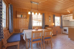 A wooden cabin interior with a dining table, chairs, a bench, a kitchen area, and a window with curtains. - Swedish House Red Water Lily (Userin)