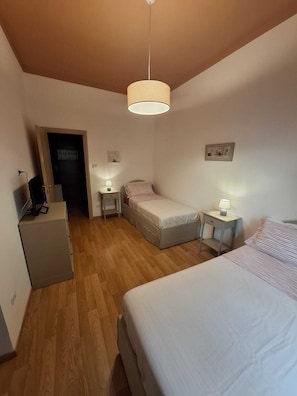 Quarto pequeno e bem iluminado, com uma cama de solteiro, mesa de cabeceira com abajur e escrivaninha com computador.