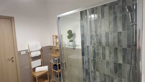 A bathroom with a glass-enclosed shower, a wooden chair, a shelf with towels, and a potted plant. - Vrbo Property (Pozzuoli)