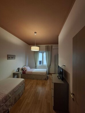 Ein kleines Hotelzimmer mit einem Einzelbett, einem Nachttisch, einem Schreibtisch mit Stuhl, einem Fernseher und einem Fenster mit Vorhängen.